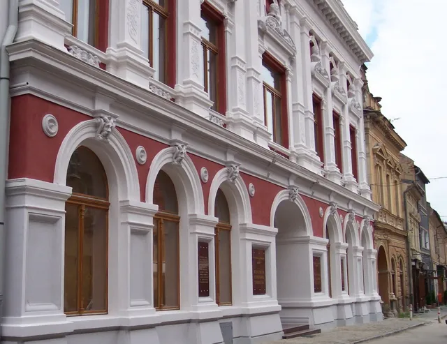 O clădire cu fațadă albă și roșie prezintă ferestre înalte, arcade și elemente decorative sculptate pe parcursul nivelurilor superioare și inferioare.