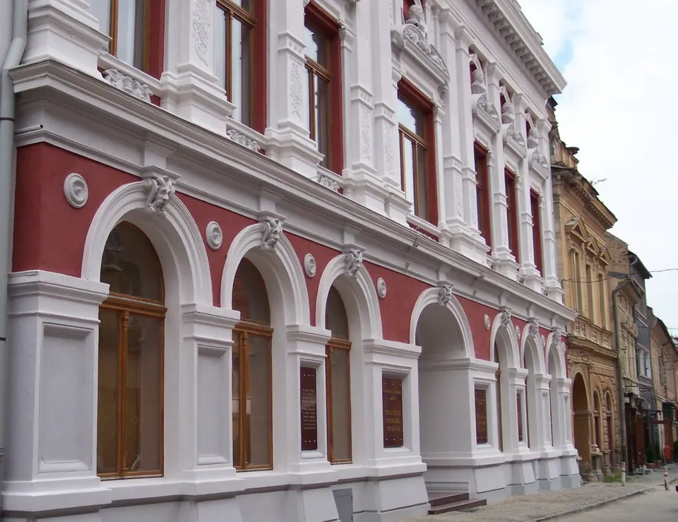 O clădire cu fațadă albă și roșie prezintă ferestre înalte, arcade și elemente decorative sculptate pe parcursul nivelurilor superioare și inferioare.
