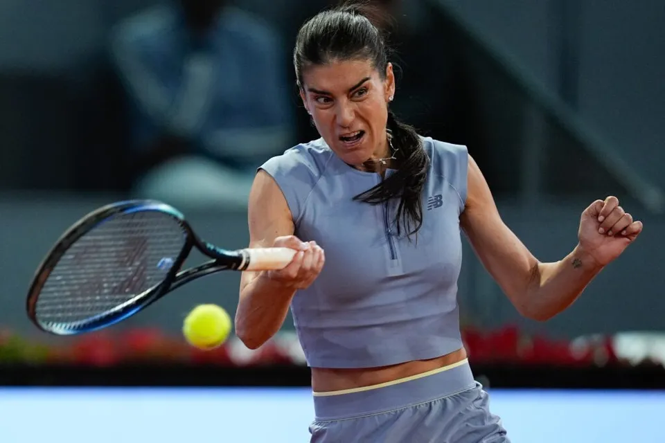 O femeie cu părul negru, purtând un tricou albastru fără mâneci, lovește o minge galbenă cu o rachetă de tenis pe un teren albastru.