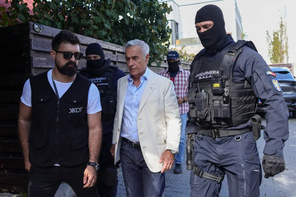 Un bărbat într-un sacou deschis merge printre trei bărbați purtând uniformă neagră și măști, unul dintre aceștia având pe vest inscripția Jandarmeria.