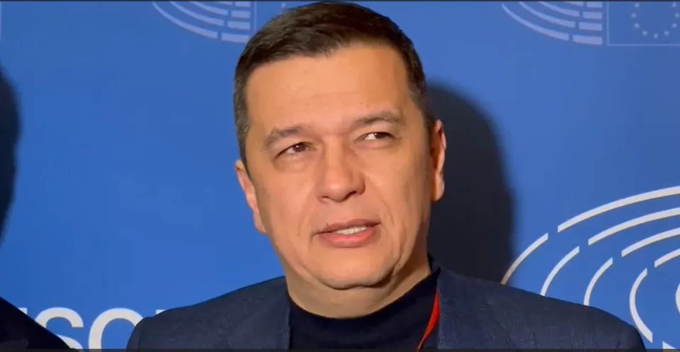 Un bărbat cu părul scurt și haine închise la culoare stă în fața unui fundal albastru cu elemente grafice albe.