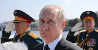 Un bărbat în costum în prim-plan este înconjurat de doi bărbați în uniforme militare care poartă căciuli și execută salutul militar.