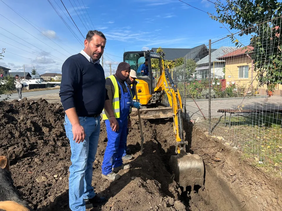Un bărbat în hanorac albastru și blugi stă lângă un lucrător cu vestă reflectorizantă și o excavatoare galbenă lângă un șanț.