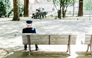 Un bărbat cu șapcă stă singur pe o bancă de lemn într-un parc, cu alți oameni așezați pe o bancă în fundal.