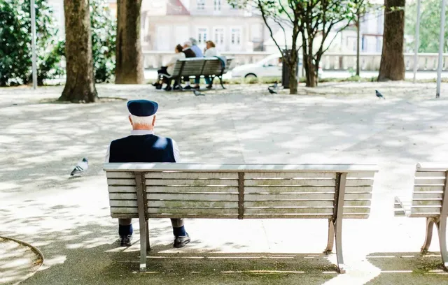 Un bărbat cu șapcă stă singur pe o bancă de lemn într-un parc, cu alți oameni așezați pe o bancă în fundal.