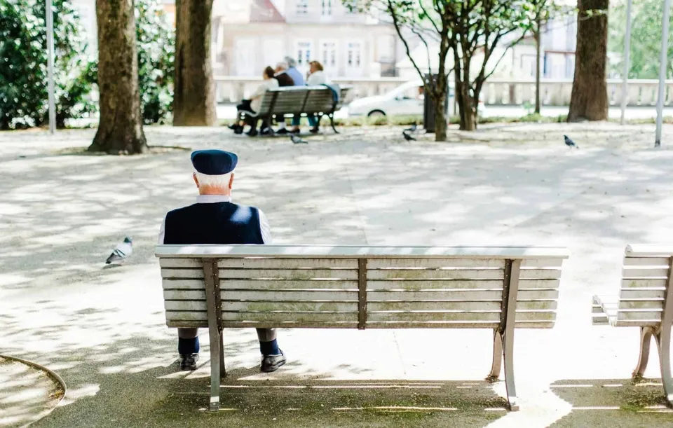 Un bărbat cu șapcă stă singur pe o bancă de lemn într-un parc, cu alți oameni așezați pe o bancă în fundal.
