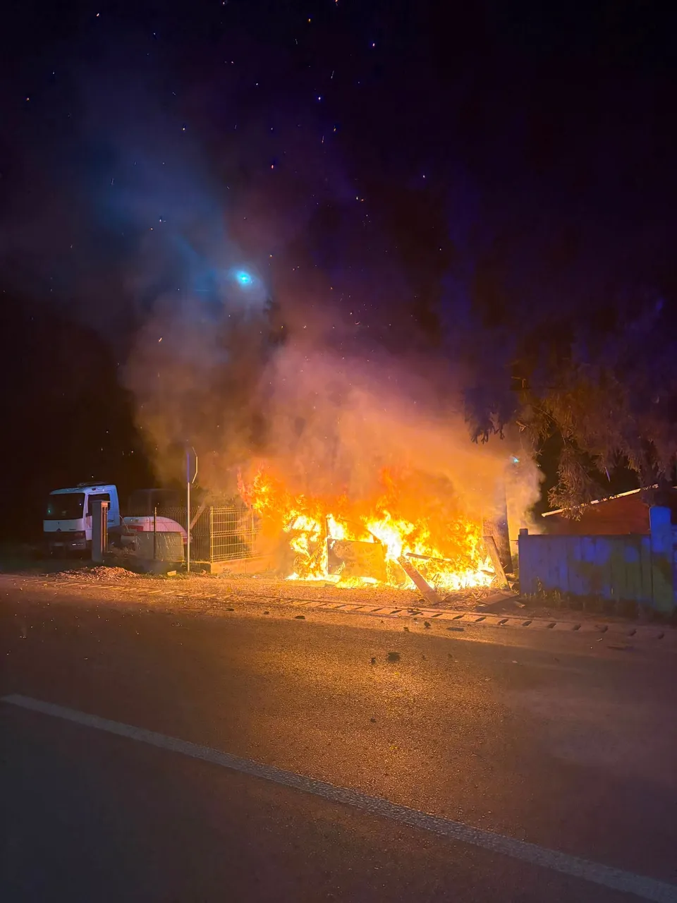 Un autoturism este acoperit de flăcări pe marginea unui drum asfaltat, lângă un gard și un van alb, sub un cer nocturn.