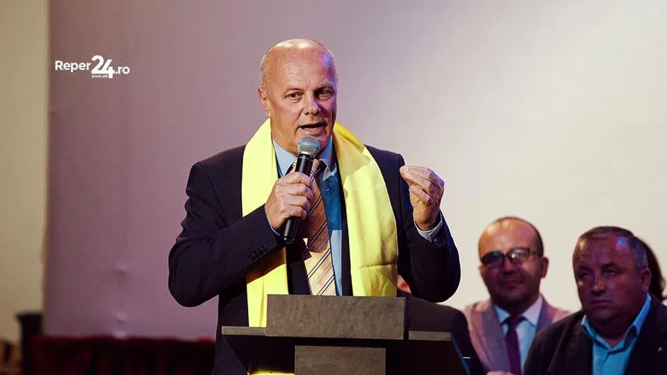 Un bărbat cu părul scurt și pielea deschisă poartă un sacou închis și un accesoriu galben, vorbind la un microfon în fața unui podium.
