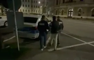 Trei bărbați stau pe un drum asfaltat lângă un autovehicul alb parcat noaptea, în fața unor clădiri iluminat.