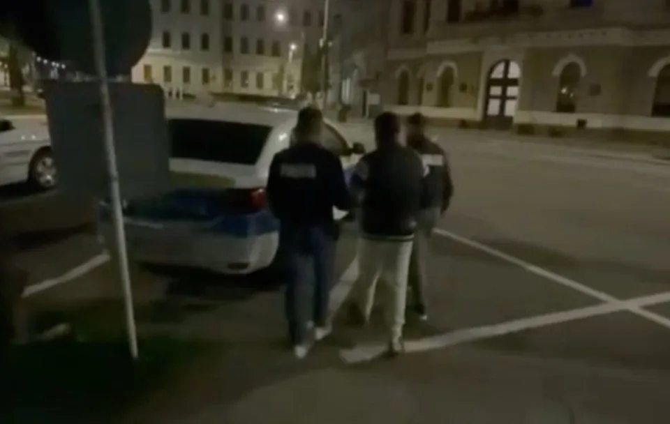 Trei bărbați stau pe un drum asfaltat lângă un autovehicul alb parcat noaptea, în fața unor clădiri iluminat.