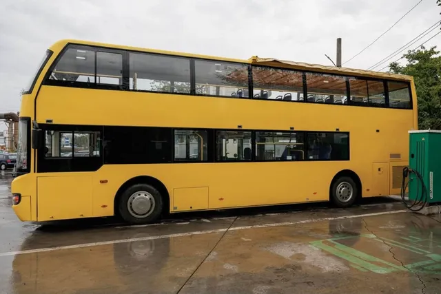 Un autobuz turistic supraetajat de culoare galbenă este parcat pe un teren asfaltat umed sub un cer înnorat.
