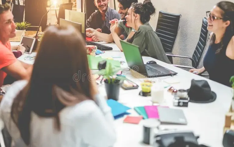 Un grup de oameni stă la o masă albă de birou cu laptopuri, telefoane și obiecte de birou în fața lor.
