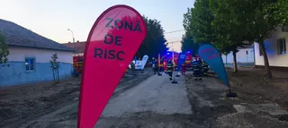 Un steag roșu cu text negru stă pe un drum de pământ lângă un vehicul de serviciu și persoane în uniforme colorate.