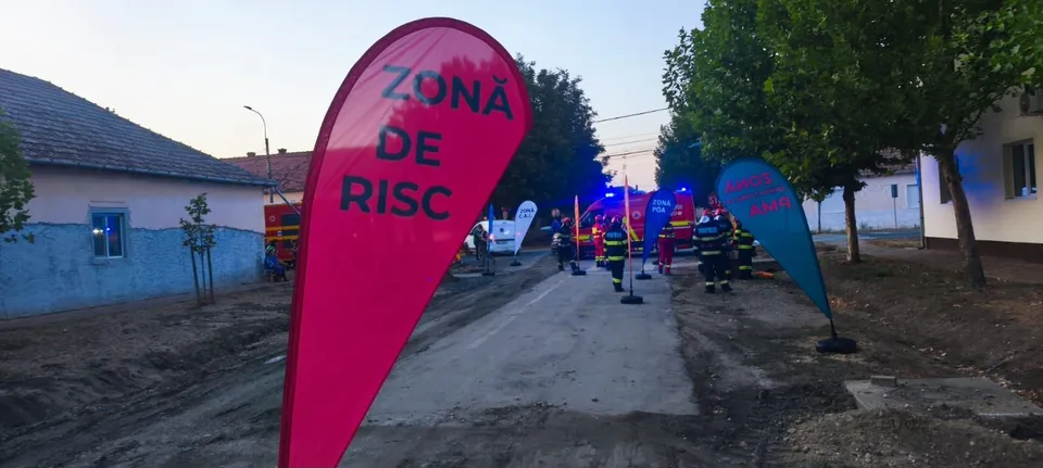 Un steag roșu cu text negru stă pe un drum de pământ lângă un vehicul de serviciu și persoane în uniforme colorate.
