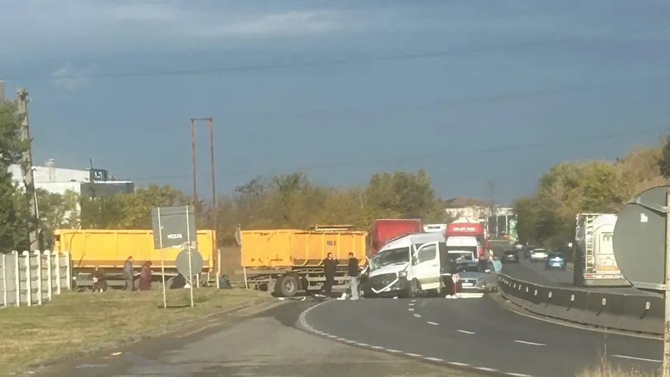 Două vehicule de marfă galbene și un camion alb sunt parcate pe marginea unui drum asfaltat lângă un gard de beton și vegetație.