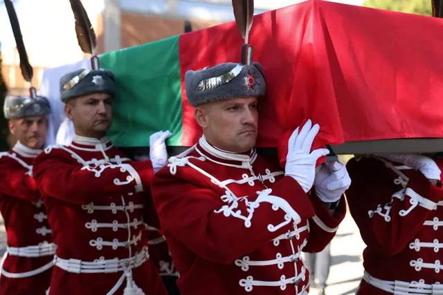 Patru bărbați în uniforme roșii cu detalii albe și pălării gri transportă un obiect acoperit cu un drapel roșu.