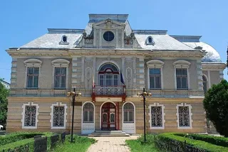 O clădire cu două etaje prezintă fațadă decorată, ferestre cu gratii și un balcon central situat deasupra unui hol de intrare cu uși duble.
