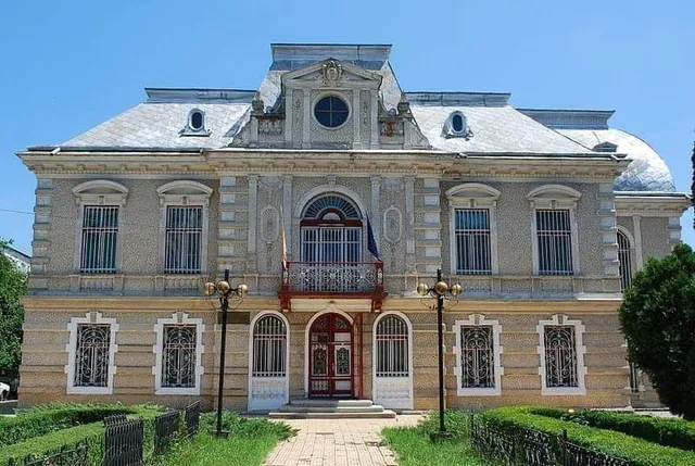 O clădire cu două etaje prezintă fațadă decorată, ferestre cu gratii și un balcon central situat deasupra unui hol de intrare cu uși duble.