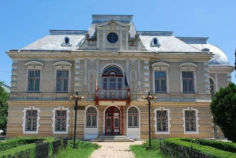 O clădire cu două etaje prezintă fațadă decorată, ferestre cu gratii și un balcon central situat deasupra unui hol de intrare cu uși duble.