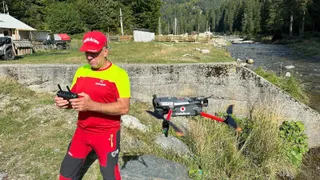 Un bărbat cu șapcă roșie și tricou galben și roșu manipulează o telecomandă lângă o dronă neagră cu marcaje roșii și albastre pe iarbă.