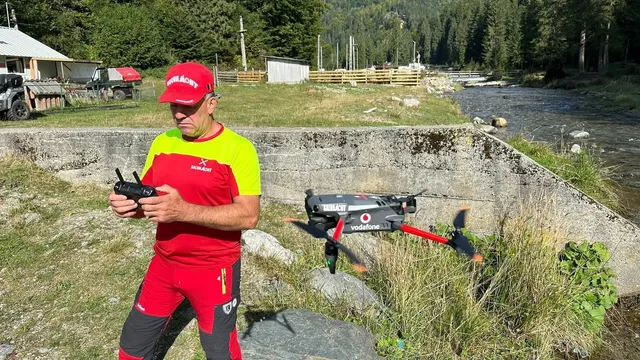 Un bărbat cu șapcă roșie și tricou galben și roșu manipulează o telecomandă lângă o dronă neagră cu marcaje roșii și albastre pe iarbă.