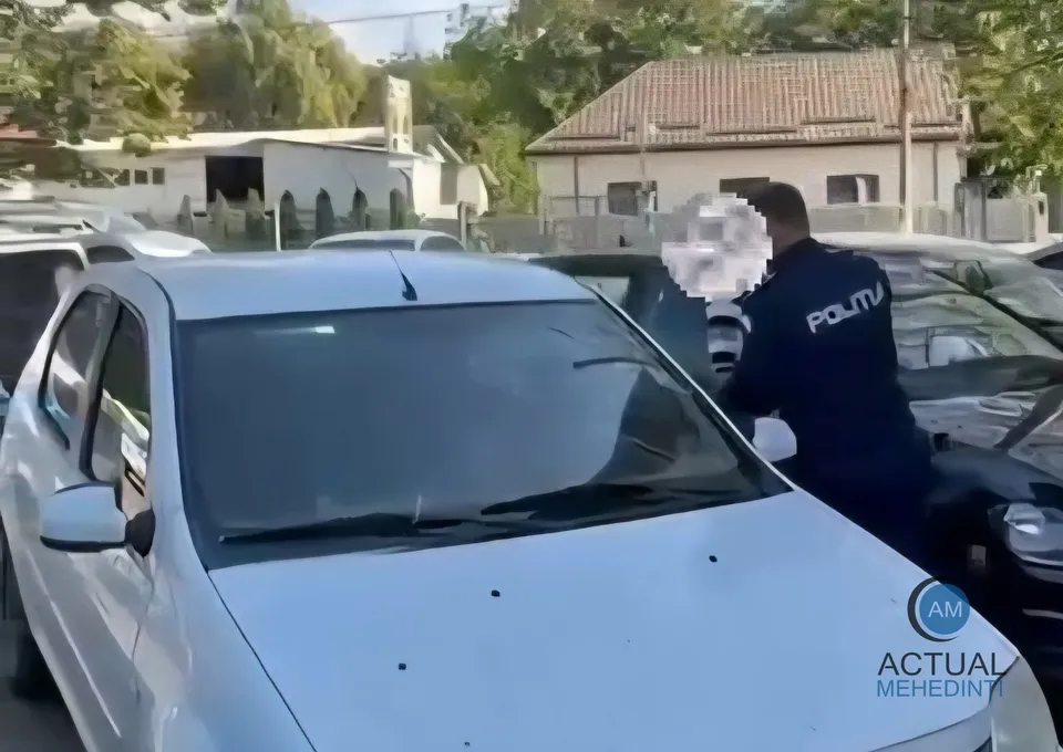 Un agent de poliție cu inscripție pe spate stă lângă o mașină albastră într-o parcare cu alte autoturisme și clădiri în fundal.
