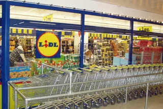 Rânduri de cărucioare de cumpărături metalice sunt așezate în fața unui magazin cu rafturi pline de diverse produse și un logo galben și roșu.