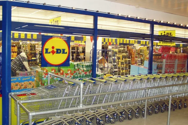 Rânduri de cărucioare de cumpărături metalice sunt așezate în fața unui magazin cu rafturi pline de diverse produse și un logo galben și roșu.