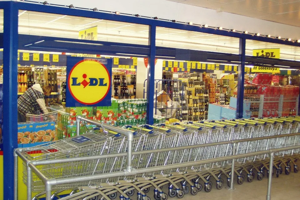 Rânduri de cărucioare de cumpărături metalice sunt așezate în fața unui magazin cu rafturi pline de diverse produse și un logo galben și roșu.