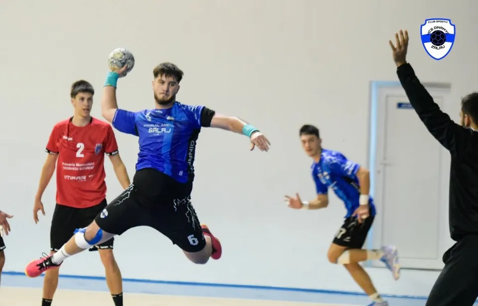 Un jucător de handbal în echipament albastru sare în aer ținând o minge, în timp ce alți jucători se află pe terenul de sport.