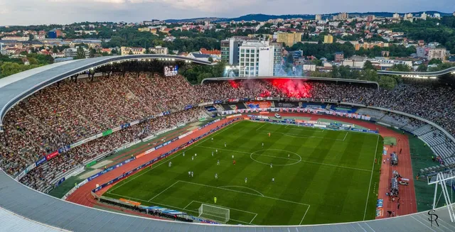 O vedere de sus a unui stadion de fotbal plin de spectatori, cu un teren verde și tribune înconjurate de clădiri și dealuri în fundal.