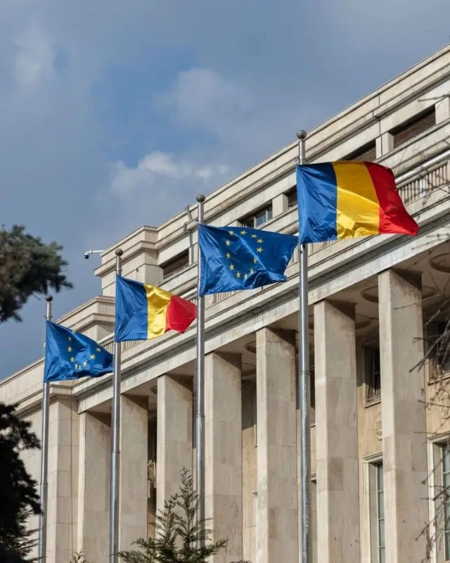 Patru steaguri, inclusiv steagurile Uniunii Europene și ale României, sunt arborate pe catarge în fața unei clădiri cu coloane de piatră sub un cer înnorat.