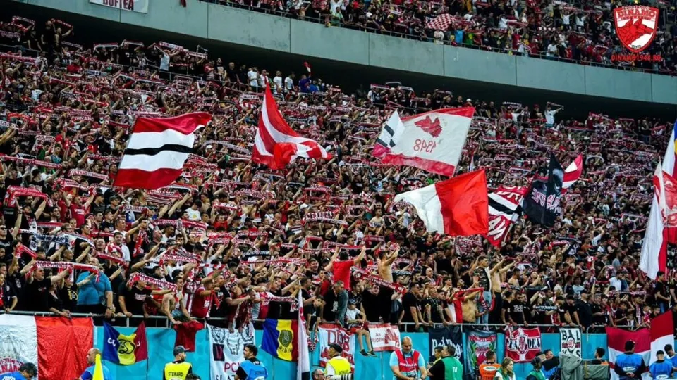 O mulțime de oameni stă pe tribunele unui stadion, ridicând diverse steaguri colorate și eșarfe în timpul unui eveniment sportiv.