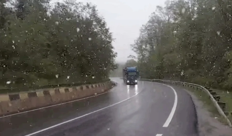 Un camion circulă pe un drum asfaltat umed, înconjurat de copaci sub un cer gri și ploaie.