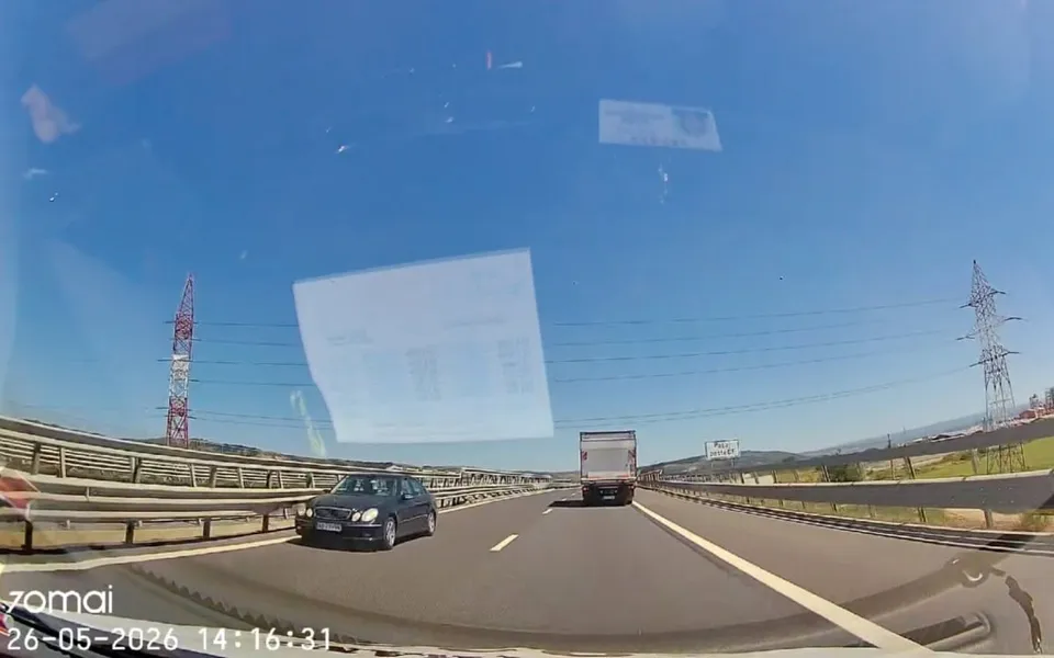 O mașină neagră și un camion alb circulă pe o autostradă sub un cer senin, lângă bariere metalice și turnuri de înaltă tensiune.