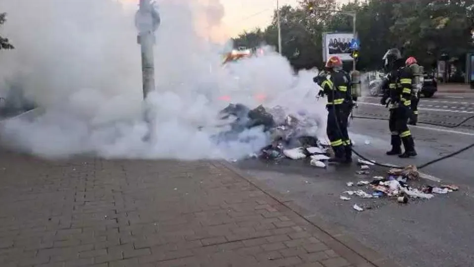 Trei pompieri în echipament de protecție portocalie și negru stau lângă un nor de fum alb pe un drum asfaltat lângă un trotuar.