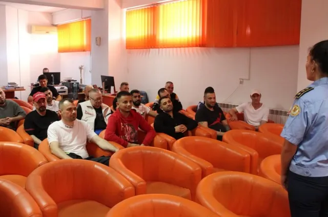 Un grup de bărbați stă așezat în scaune de culoarea portocalie într-o încăpere cu ferestre acoperite de draperii portocalii, în fața unui uniformat.