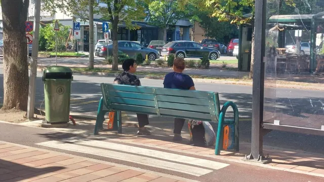 Două persoane stau pe o bancă verde de lângă un coș de gunoi verde, pe un trotuar lângă o pistă de biciclete.