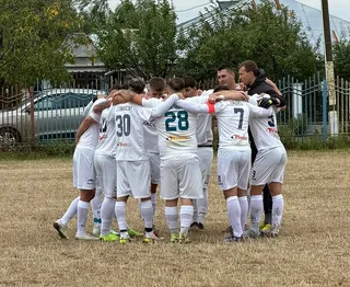 Un grup de fotbaliști îmbrăcați în echipamente albe stau într-un cerc, cu brațele pe umerii colegilor, pe un teren de iarbă uscată.