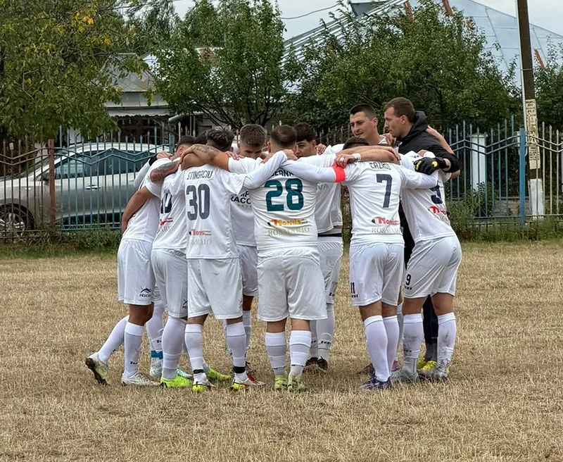 Un grup de fotbaliști îmbrăcați în echipamente albe stau într-un cerc, cu brațele pe umerii colegilor, pe un teren de iarbă uscată.