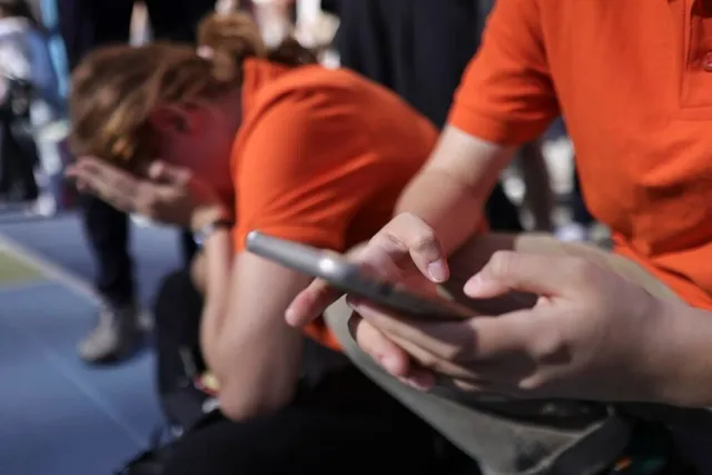 O persoană în tricou portocaliu ține un telefon mobil, în timp ce în fundal o altă persoană stă aplecată cu fața acoperită de mâini.