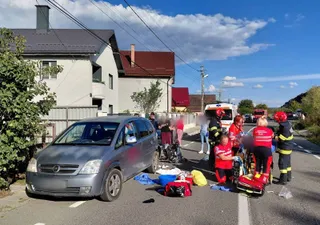 Echipe medicale în uniforme roșii și negre stau pe asfalt lângă o mașină gri și echipamente medicale într-o zonă rezidențială.