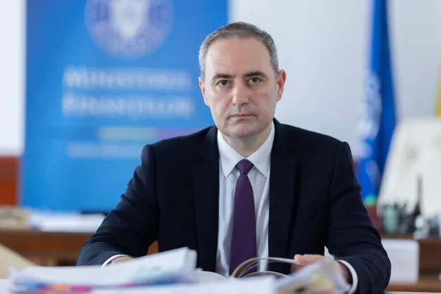 Un bărbat cu părul scurt, purtând sacou negru, cămașă albă și cravată mov, stă la o masă în fața unui fundal albastru.