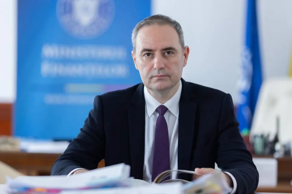 Un bărbat cu părul scurt, purtând sacou negru, cămașă albă și cravată mov, stă la o masă în fața unui fundal albastru.