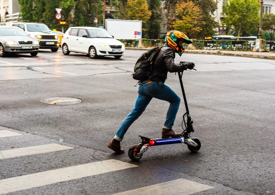 Un bărbat cu cască și rucsac poartă o trotinetă electrică pe o trecere de pietoni într-o intersecție cu mașini în fundal.