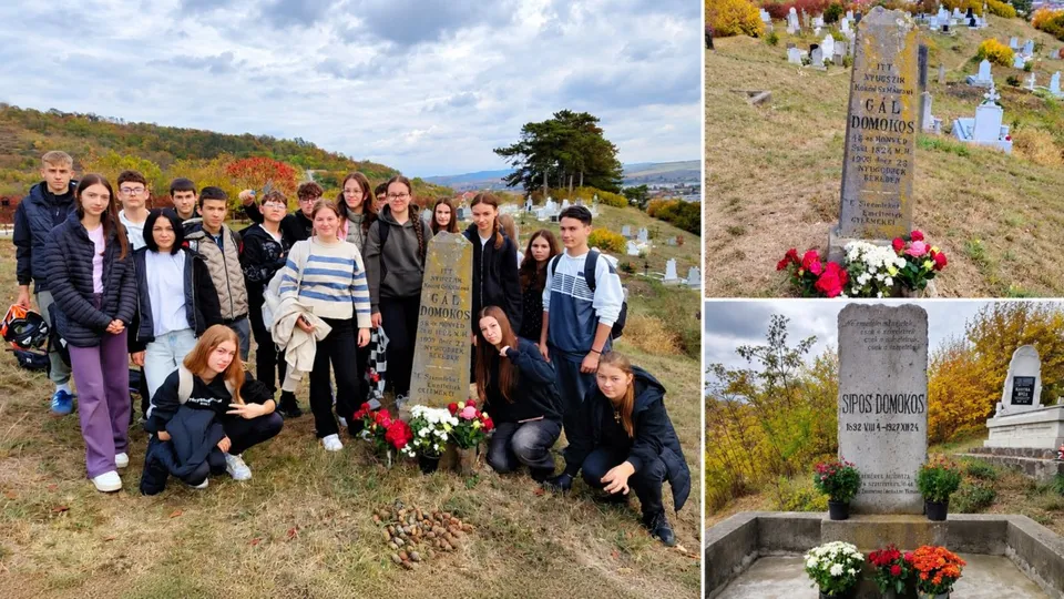 Un grup de elevi stă lângă două morminte înconjurate de flori într-un cimitir aflat pe un teren înclinat sub un cer înnorat.