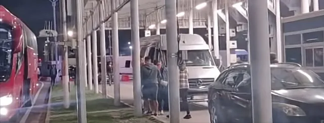 Autobuzul alb și mașinile sunt parcate sub o structură acoperită de coloane, lângă un autocar roșu, în timpul nopții.
