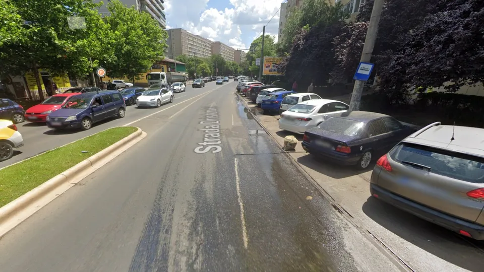 O stradă asfaltată cu mașini parcate pe margini, linii de marcaj rutiere și vegetație pe partea stângă sub un cer cu nori.