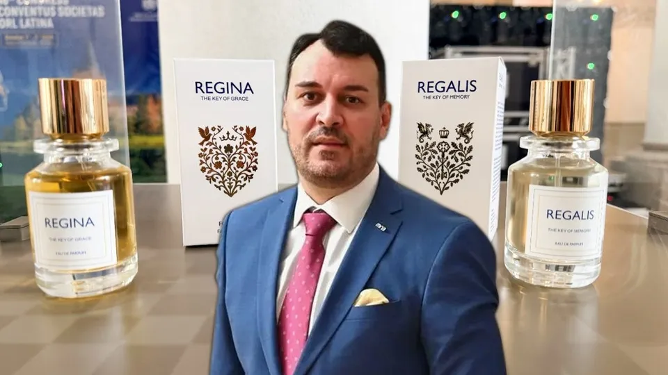 Un bărbat într-un costum albastru și cravată roz stă între două flacoane de parfum și două cutii albe pe o suprafață lucioasă.