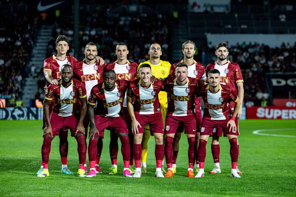 Un grup de unsprezece jucători de fotbal în echipamente roșu și alb stau posați pe un teren de fotbal sub luminile unui stadion.